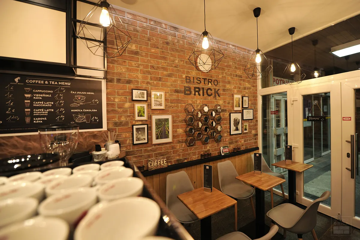 Tehlová stena s nápisom BISTRO BRICK, rámovanými fotografiami a drevenými stolmi so sivými stoličkami.