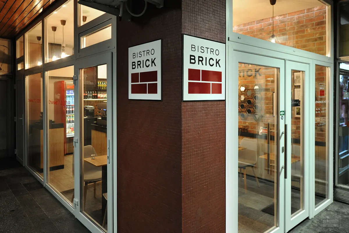 Presklený vstup do bistra Bistro Brick s tmavým obkladom a výkladom, cez ktorý je vidno interiér s tehlovou stenou.