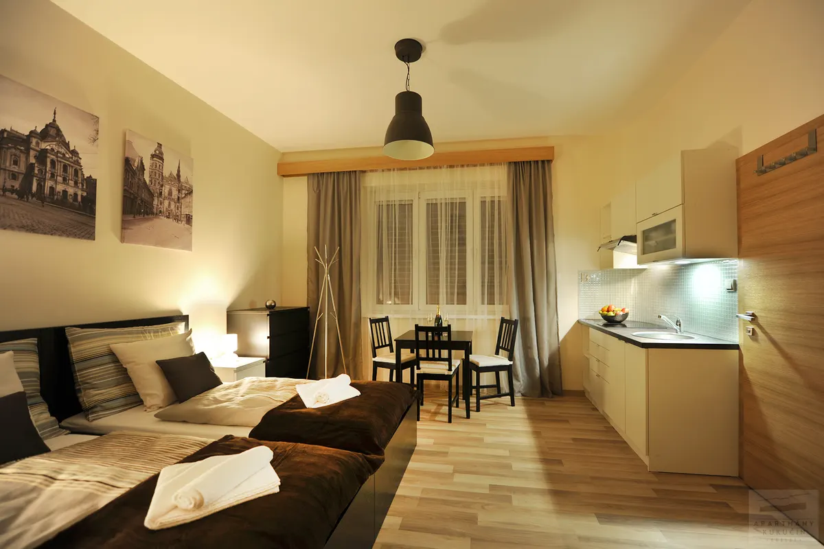 Apartmán s posteľou, jedálenským stolíkom pri okne a bielou kuchynskou linkou pri stene.