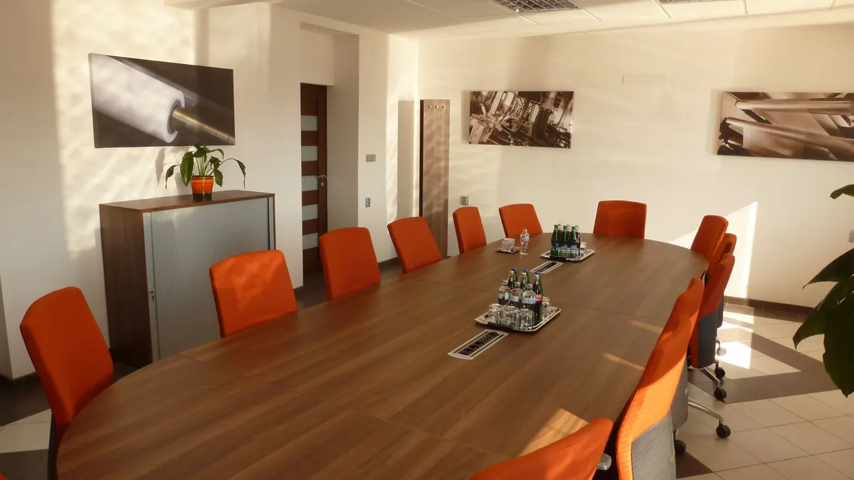 Rokovacia miestnosť s dlhým dreveným stolom, oranžovými stoličkami a bielymi stenami.